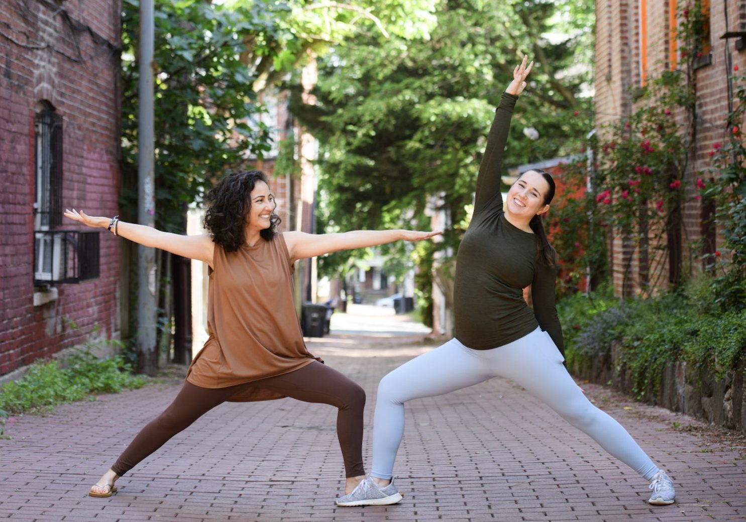 Kids yoga teacher Ferdaouis and staff member Kimberly smiling in warrior yoga poses in a brick-lined alley with vibrant green trees in the background